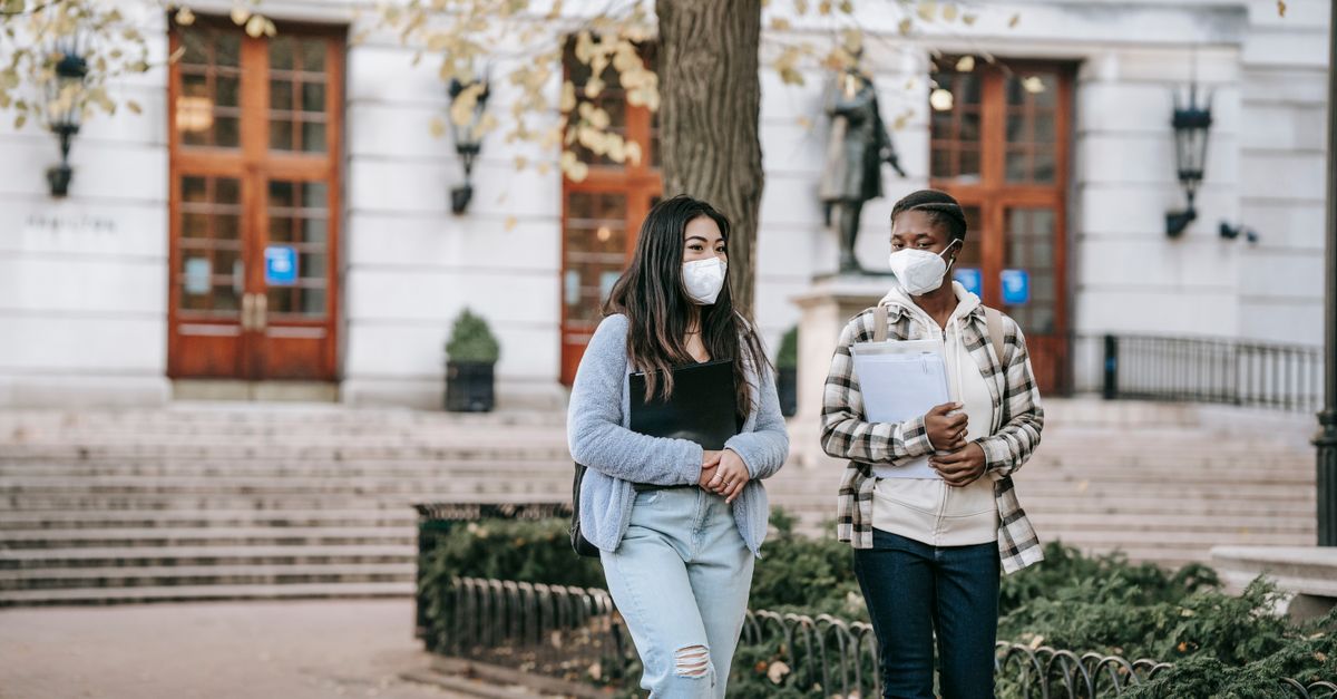 Focused young multiracial female students in casual clothes and protective masks chatting and carrying folders while strolling on campus sidewalk together